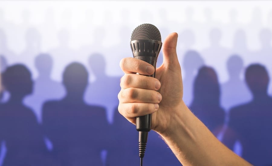 Hand holding microphone and showing thumbs up in front of a crowd of silhouette people. Public speaking and giving speech