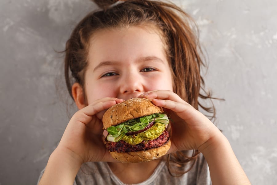 Beautiful happy hungry baby girl eating vegan burger.  Vegan beet chickpea burgers with vegetables, guacamole and rye buns. Healthy child vegan food concept.